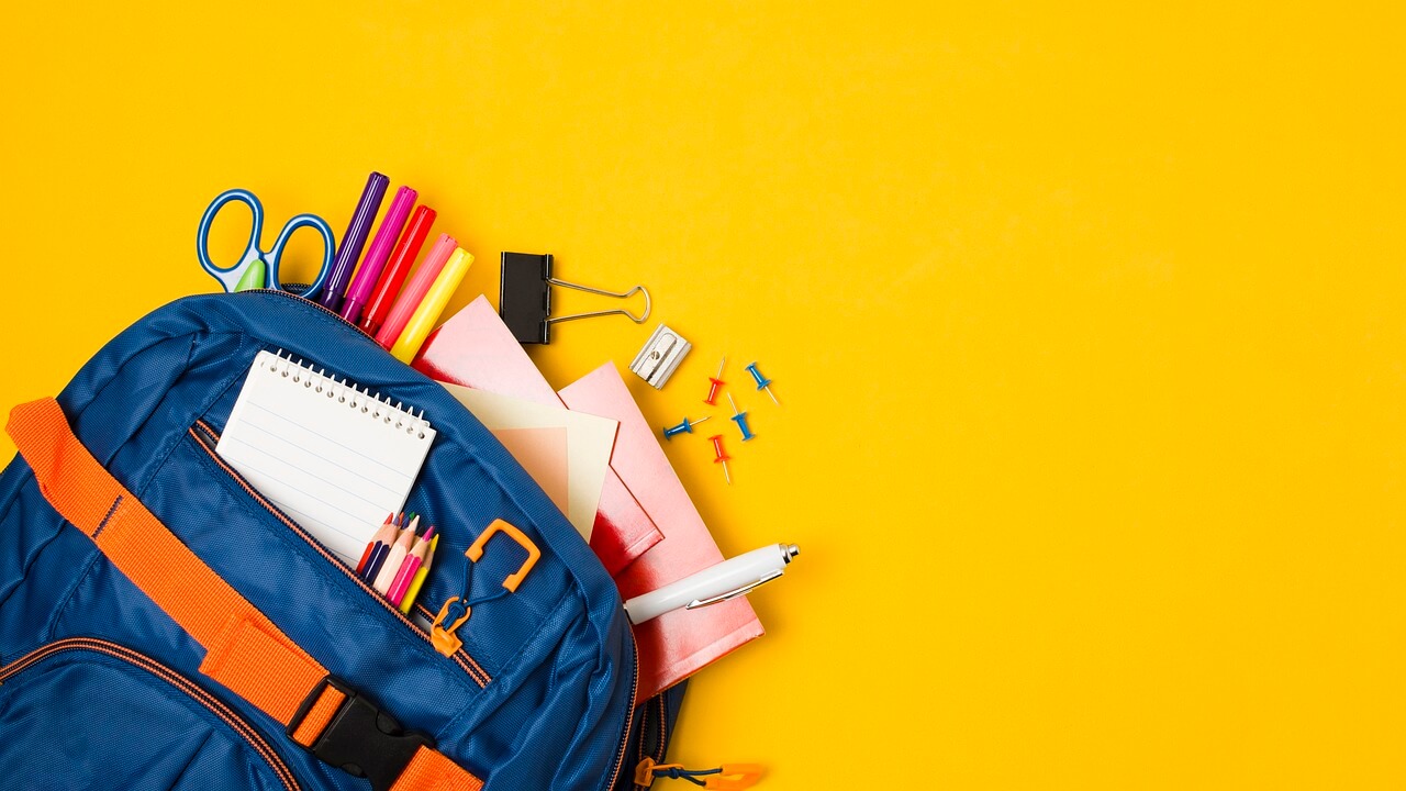 Cartable bleu ouvert sur fond uni jaune. La cartable laisse dépasser un calepin, des ciseaux et des crayons...