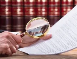 photo d'une main qui tient une loupe d'un coté et un contrat de l'autre. En arrière plan on découvre des livres